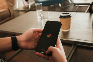 The image captures a casual moment where a person is holding a smartphone with the Instagram logo displayed on the screen. The background shows a blurred setting of a group discussion or meeting in a cozy indoor environment. A coffee cup and glasses of water are placed on the table, adding to the relaxed atmosphere. The focus on the phone contrasts with the busy backdrop, highlighting themes of digital connection amidst real-life interactions. The image captures a casual moment where a person is holding a smartphone with the Instagram logo displayed on the screen. The background shows a blurred setting of a group discussion or meeting in a cozy indoor environment. A coffee cup and glasses of water are placed on the table, adding to the relaxed atmosphere. The focus on the phone contrasts with the busy backdrop, highlighting themes of digital connection amidst real-life interactions.