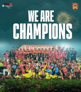 Football team posing with the trophy on the pitch under a "We Are Champions" graphic, with fireworks in the night sky above.