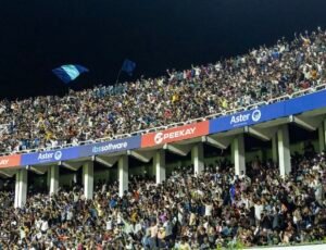 Packed stadium stands at night at a SuperLeagueKerala match, with thousands of fans cheering, raising their arms and lighting up phone flashlights while two blue flags wave above the crowd."