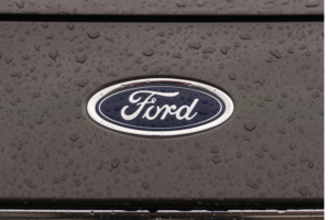 Close-up view of a Ford car logo on a wet hood, with raindrops covering the surface. Close-up view of a Ford car logo on a wet hood, with raindrops covering the surface.