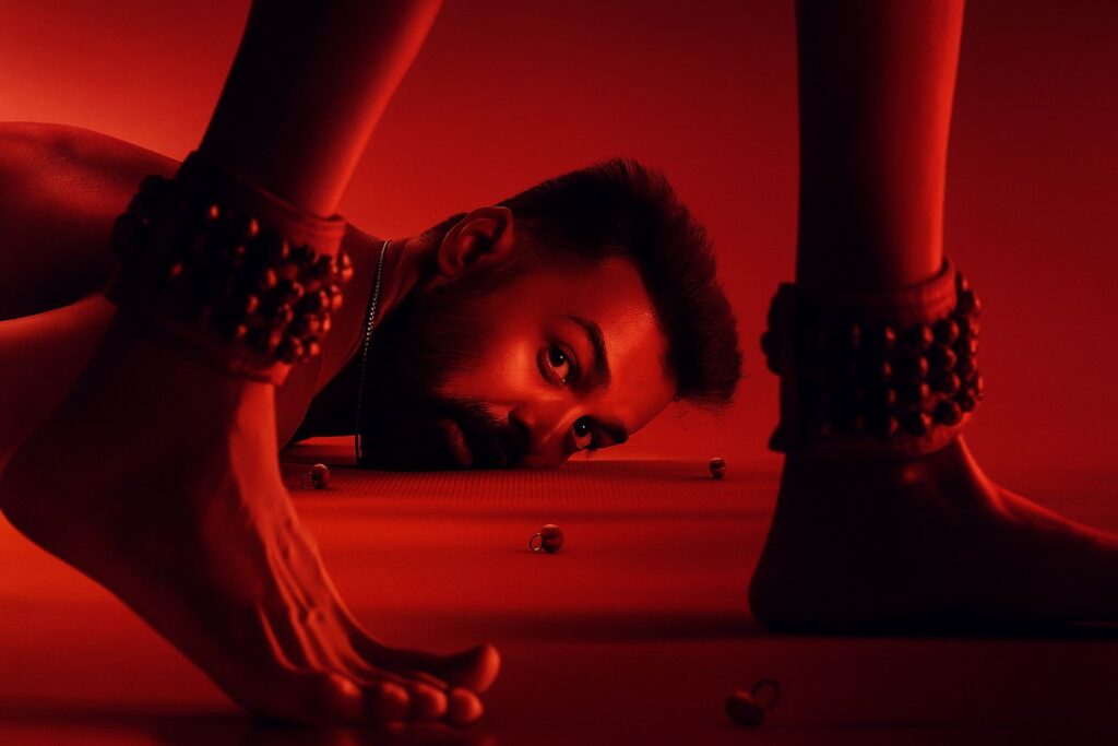 A man lies on the floor under dramatic red lighting, gazing intensely ahead, framed between two feet adorned with traditional anklets. Small bells are scattered on the ground, creating a powerful and artistic visual composition. the film dies irea