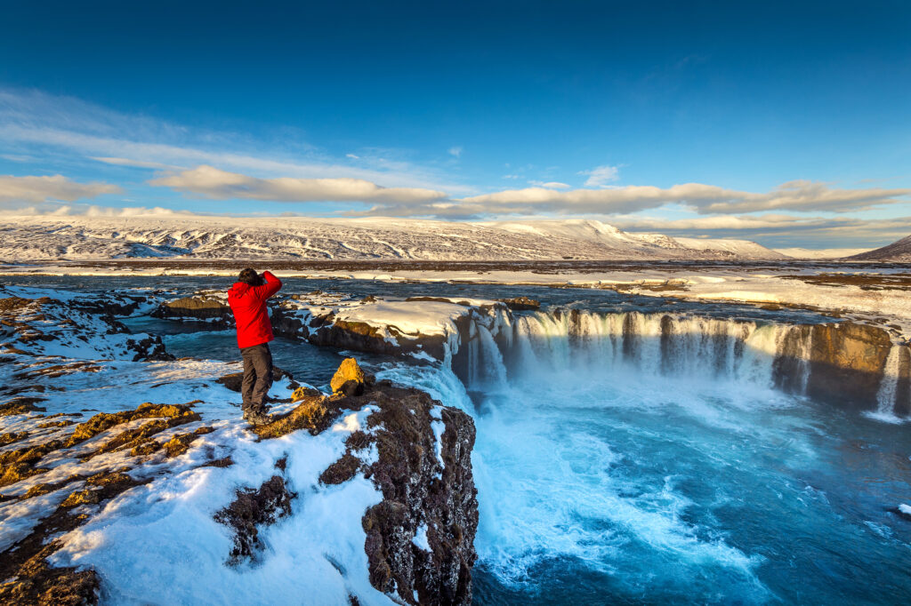 A traveler in a red jacket captures the stunning frozen landscape and waterfall under a bright blue sky, showcasing Iceland winter magic at its best.
