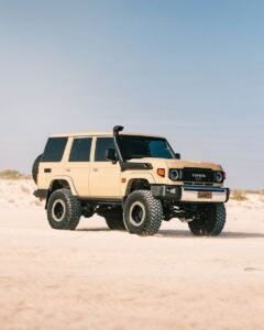 Toyota Land Cruiser SUV with large off-road tires parked on a sandy desert landscape under a clear blue sky.