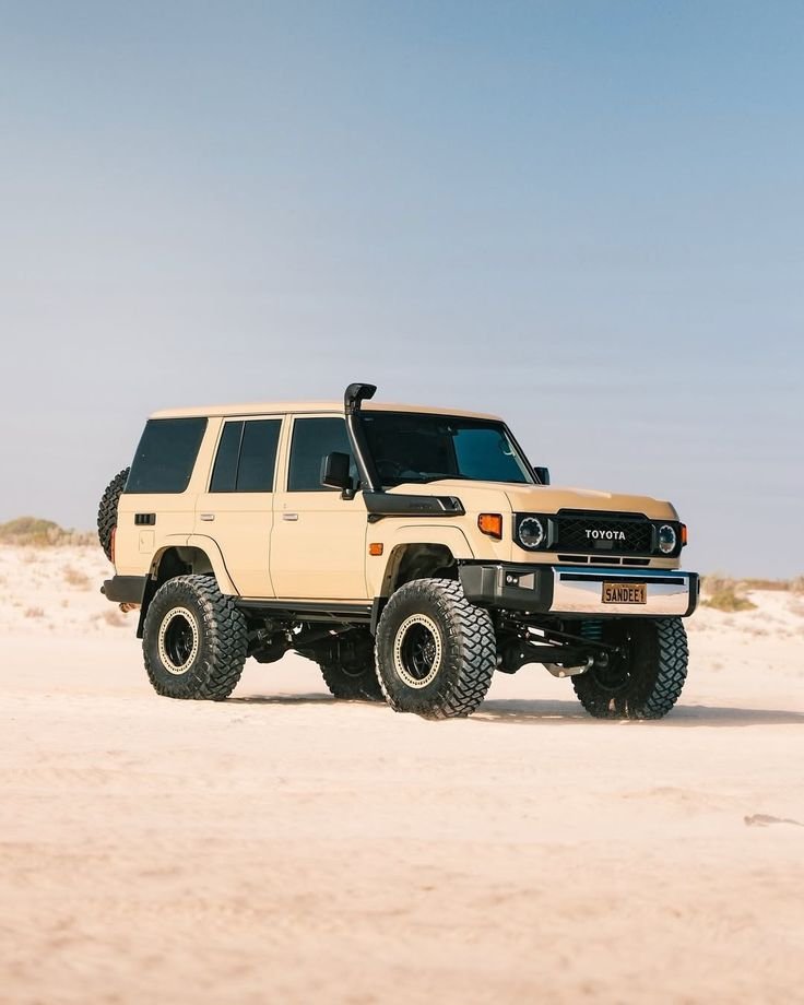 Toyota Land Cruiser SUV with large off-road tires parked on a sandy desert landscape under a clear blue sky.